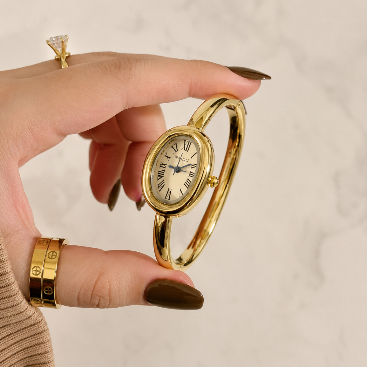 A gold bangle watch with an oval face and Roman numerals, held by a hand with dark brown manicured nails against a light marble background.