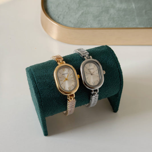 Two small oval wristwatches displayed on a dark green velvet jewelry stand. One watch has a gold-tone case with a silver-tone bracelet, and the other has a silver-tone case with a gold-tone bracelet.