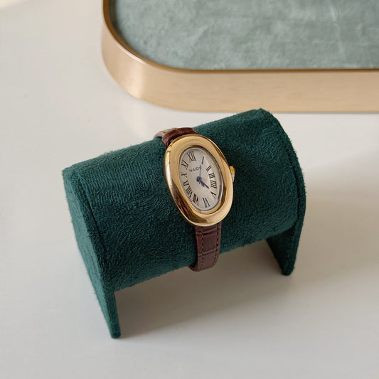 An oval wristwatch with a gold-tone case, white dial, and brown leather strap, displayed on a dark green velvet stand on a light surface.