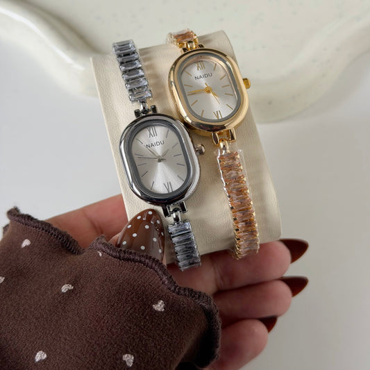 Two NAIDU brand oval watch bracelets displayed on a cream-colored stand, held by a hand wearing a brown polka-dot sleeve. One watch has a silver-toned case and bracelet with a white dial, and the other has a gold-toned case and a translucent rose-gold textured bracelet with a white dial.