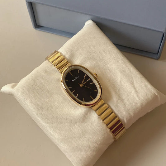 A gold-tone Hagen watch with an oval black dial and matching gold-tone link bracelet, displayed on a cream-colored cushion with its blue-gray presentation box in the background.