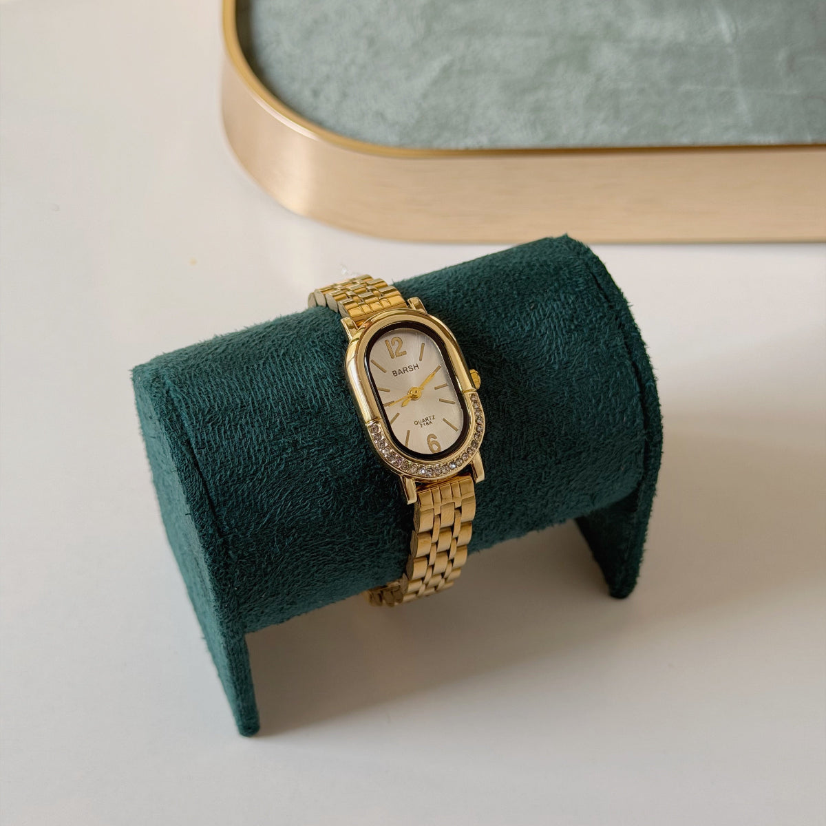 A gold-tone oval watch with a white dial and crystal-encrusted bezel, displayed on a dark green velvet watch stand against a light gray background.