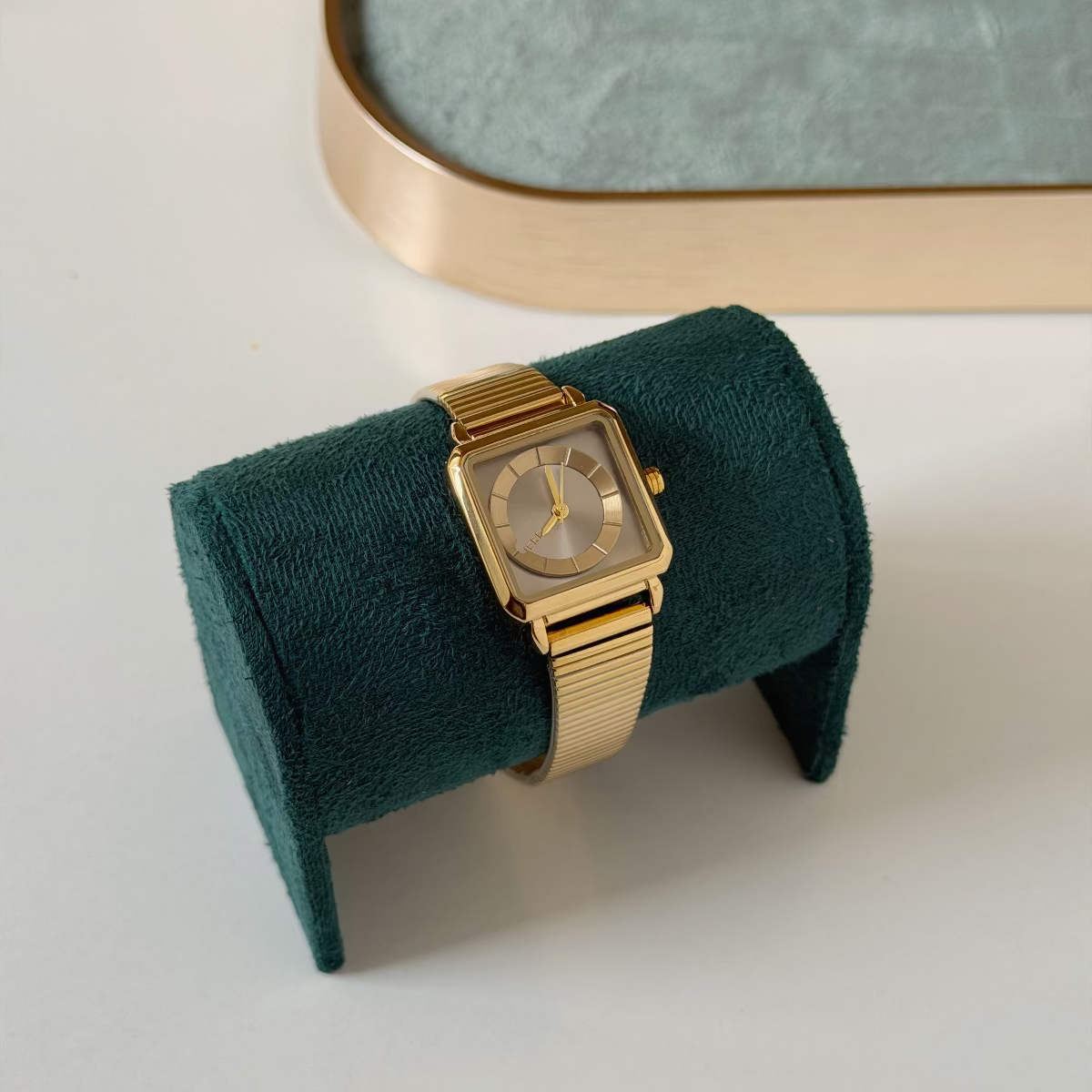 A gold-tone rectangular watch with a matching metal mesh band, displayed on a dark teal velvet watch stand against a light gray background.