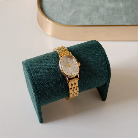 A gold-tone wristwatch with a round white dial and matching gold bracelet, displayed on a dark green velvet jewelry stand on a light surface.