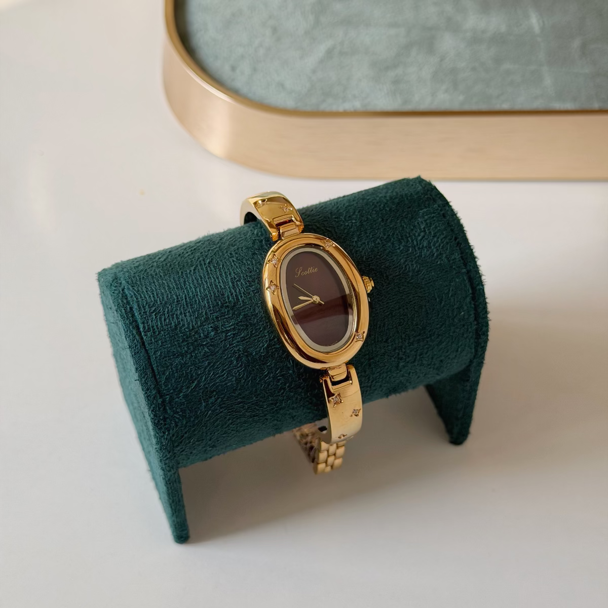 Gold-tone oval wristwatch with a dark brown dial, displayed on a dark green velvet watch stand against a white background with a blurred green jewelry box in the upper background.