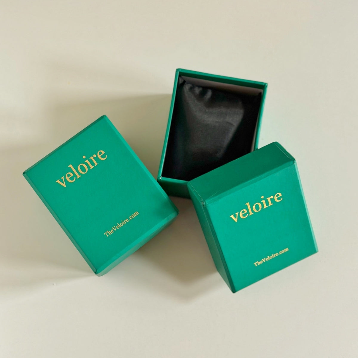 Three teal jewelry gift boxes branded 'veloire' with gold lettering, including one open box showing a black interior, arranged on a light beige background.