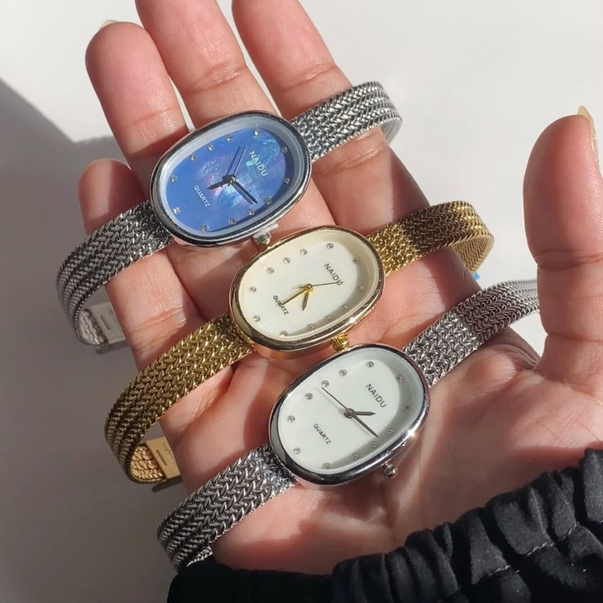 Three analog mesh-strand wristwatches are held in a person's hand. The watches have oval faces with silver-toned metal cases and bands, one with a blue dial, one with a white dial, and one with a gold-toned dial.