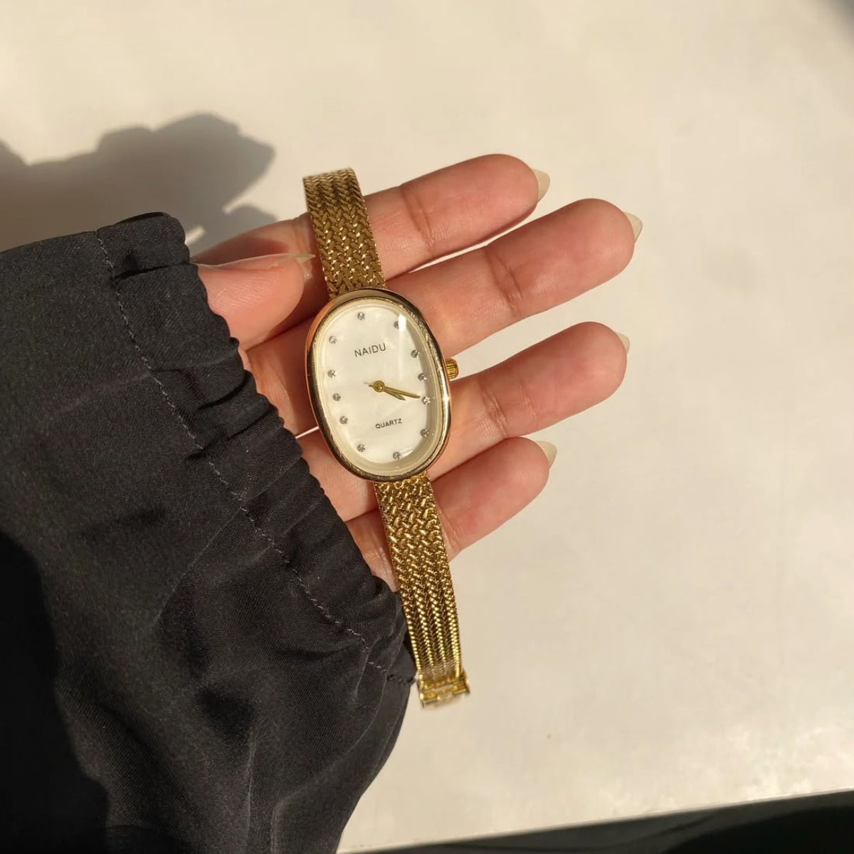 A gold-tone oval watch with a white dial and mesh bracelet, held in a person's hand against a light off-white background.