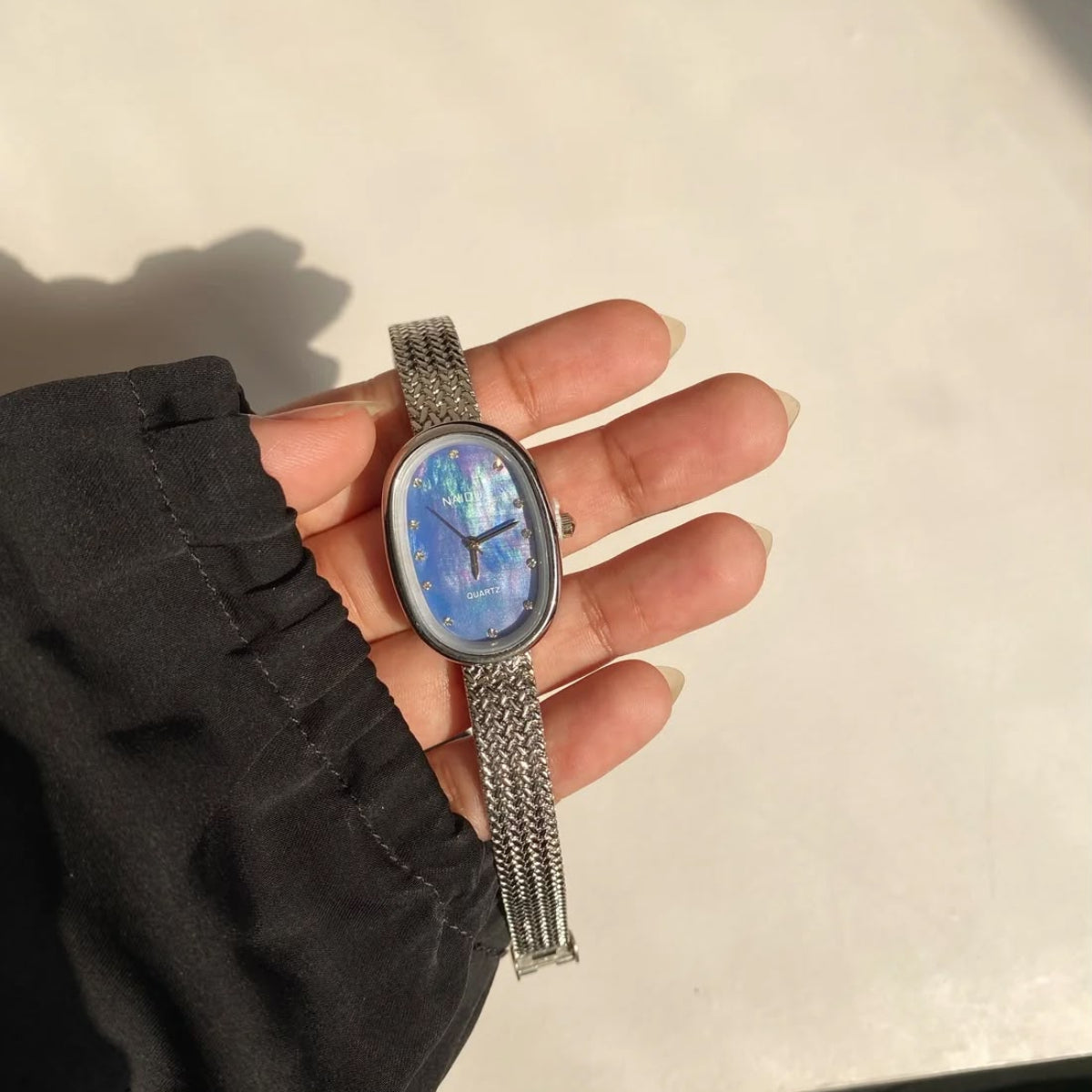 A silver mesh-strand watch with an oval mother-of-pearl blue dial, held in a person's hand wearing a black sleeve.
