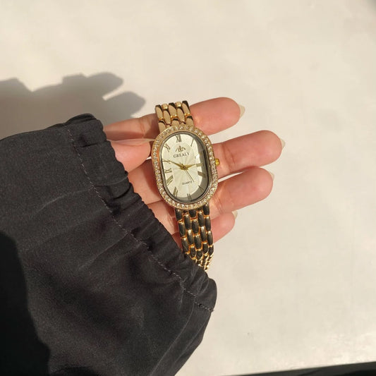 A gold-tone watch with a white dial and a gold link bracelet, held in a person's hand wearing a black sleeve.