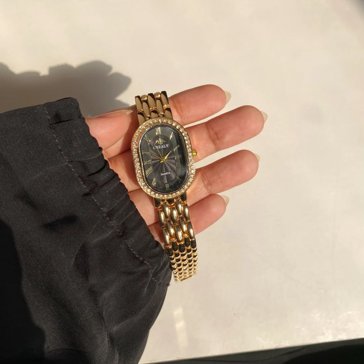 A gold-tone watch with a dark dial and crystal-embellished bezel, held in a person's hand wearing a black sleeve.