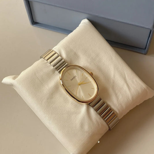 A women's oval watch by MISO, featuring a gold-tone case, gold dial, and a silver-tone link bracelet, displayed on a cream-colored fabric cushion with its blue-gray gift box in the background.